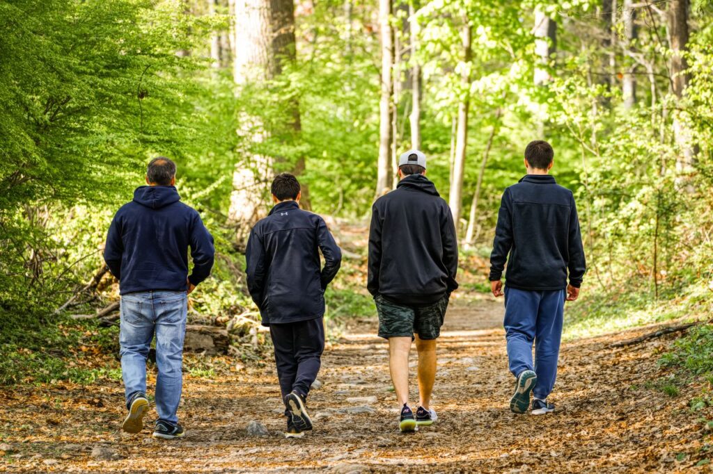 Four men in casual attire walking on a sunlit forest trail, enjoying nature.